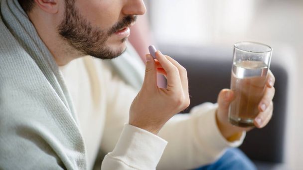 Cropped of bearded man holding a pill between his fingers, ready to swallow it with the aid of a full glass of water, highlighting a moment of personal health care - Valokuva, kuva