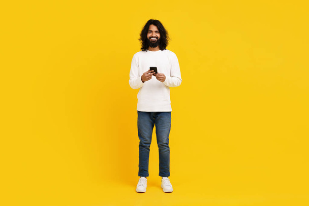Um jovem índio alegre com cabelos longos e barba fica confiante contra um cenário amarelo brilhante, segurando um smartphone em suas mãos. - Foto, Imagem
