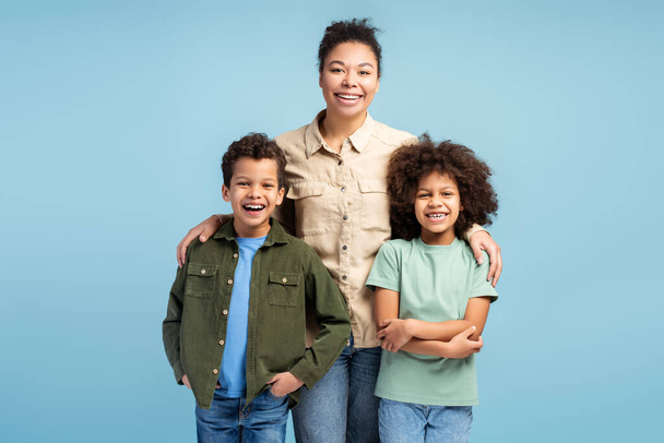Happy African American mother, little daughter and son hugging, looking at camera, standing isolated on blue background. Concept of motherhood, relationships - Photo, Image