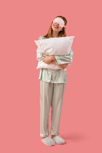 Portret van mooie jonge vrouw in pyjama met slaapmasker en kussen op roze achtergrond - Foto, afbeelding