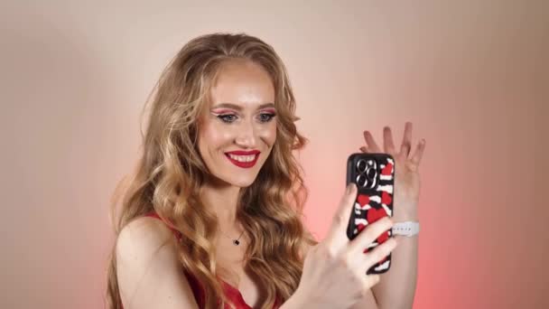Une jeune femme en robe rouge prend un selfie avec de longs cheveux blonds et du maquillage. La fille sourit et regarde la caméra en gros plan - Séquence, vidéo