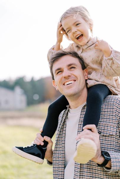 Een lachend meisje doet haar haar recht terwijl ze op de schouders van een lachende vader zit. Hoge kwaliteit foto - Foto, afbeelding