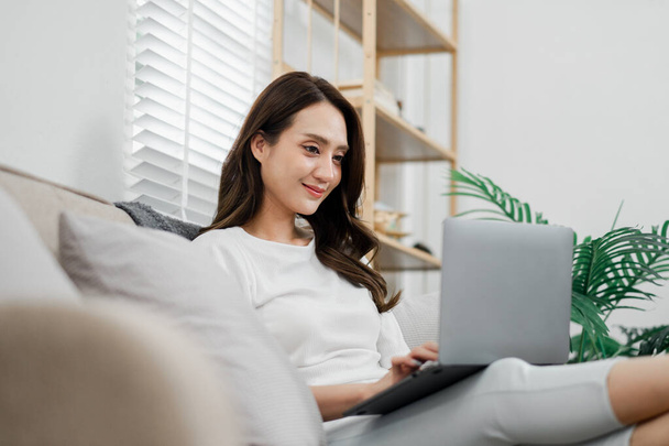 Relaxed woman enjoys working from home, comfortably lounging on a couch with her laptop in a well-lit living space. - Photo, Image