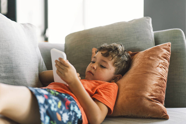 Un niño con el pelo rizado está reclinado cómodamente en un sofá gris con almohadas, profundamente inmerso en el juego en el teléfono. pausado día en el interior. - Foto, Imagen