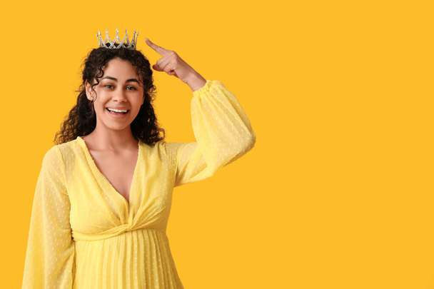 Beautiful young African-American woman in stylish prom dress pointing at tiara on yellow background - Photo, Image