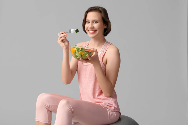 Belle jeune femme heureuse avec boule de fitness et bol de salade fraîche sur fond gris. Concept de perte de poids - Photo, image