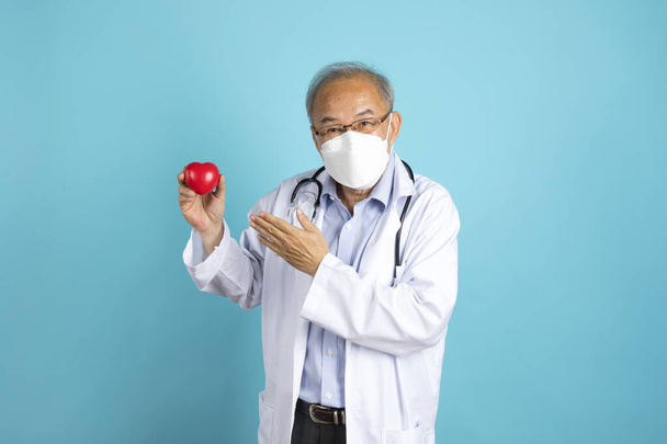 asiatico anziano medico in medico maschera e bianco cappotto mostrando rosso cuore mock fino posa su blu sfondo - Foto, immagini