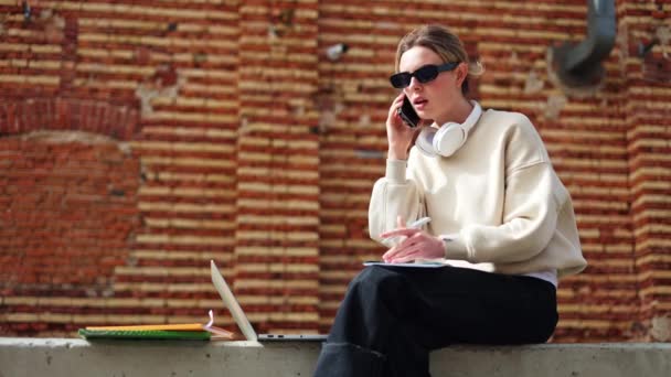 Güzel sarışın beyaz kadın, açık havada otururken modern akıllı telefon konuşmalarına dalmış. Güzel bayan temiz hava hakkında aktif konuşma sırasında titizlikle not alıyor.. - Video, Çekim