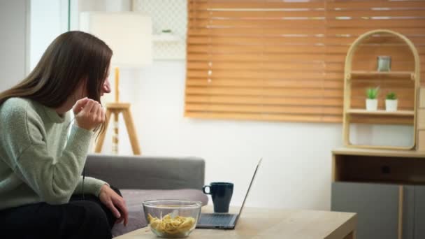 Kaunis nuori nainen syö evästeitä puhuessaan videopuhelu tyttö ystävä kautta kannettava tietokone kotona. - Materiaali, video