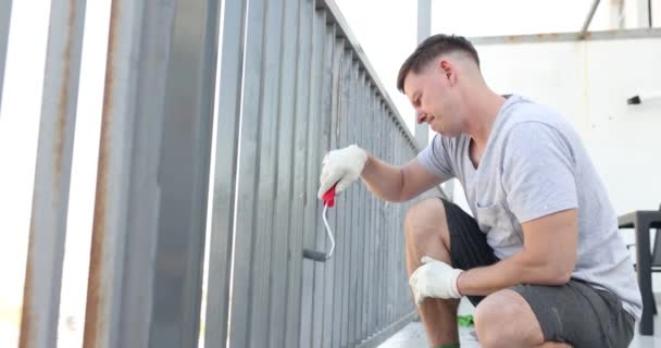 Ο άνθρωπος εφαρμόζει φρέσκο χρώμα σε σκουριασμένο κιγκλίδωμα μπαλκονιού. Ζωγραφική βεράντα καλοκαίρι - Πλάνα, βίντεο