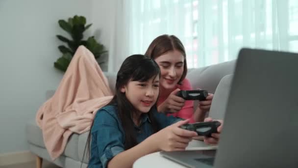 Ευτυχισμένο κορίτσι που παίζει παιχνίδι και κάθεται ενώ η μαμά ξαπλώνει στον καναπέ με τη χρήση οθόνης laptop. Καυκάσιος γονέας και ελκυστική κοπέλα περνούν χρόνο μαζί, ενώ κρατώντας joystick και να επικεντρωθεί στην νίκη παιχνίδια. Παιδαγωγική. - Πλάνα, βίντεο
