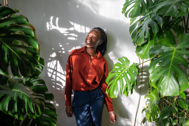 Расслабленная афроамериканка стоит в саду, наслаждаясь комнатными растениями. Беззаботная черная женщина, окруженная комнатными растениями, смотрит вдаль. Любители растений, психическое здоровье, медленная жизнь.  - Фото, изображение