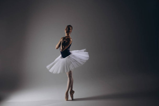 Młoda balerina w eleganckiej białej tutu i szpiczaste buty tańczące na ciemnym tle. Pełna wdzięku baletnica występująca w ciemności. Sztuka baletowa, wdzięk, elastyczność, ruch. - Zdjęcie, obraz