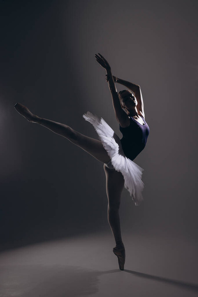 Молодая балерина в элегантной белой пачке и пуантах, исполняющая арабскую позу на тёмном фоне. Изящная балерина, выступающая в темноте. Искусство балета, грация, гибкость, движение. - Фото, изображение