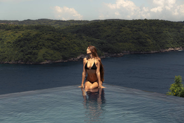 Žena v černých bikinách má klidný výhled z nekonečného bazénu s výhledem na bujnou pobřežní krajinu - Fotografie, Obrázek