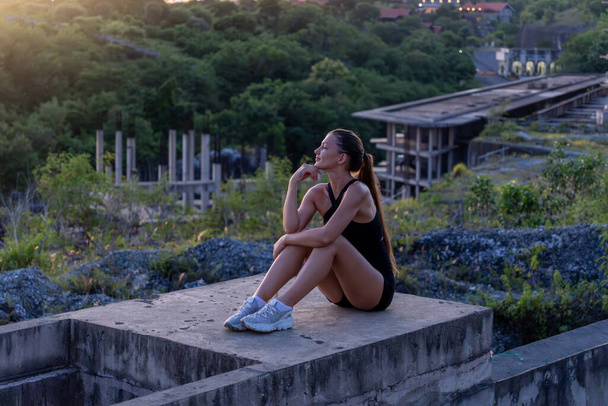 Μια νεαρή γυναίκα με αθλητική ενδυμασία κάθεται σκεπτικά σε ένα τσιμεντένιο περβάζι, με ένα εγκαταλελειμμένο κτίριο και πλούσιο πράσινο στο βάθος το σούρουπο. - Φωτογραφία, εικόνα