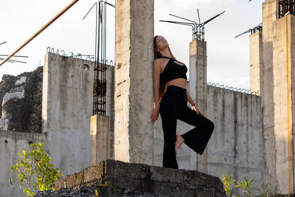 Eine Frau in schwarzer Freizeitkleidung steht kontemplativ an Betonpfeilern und Balken vor klarem Himmel. - Foto, Bild