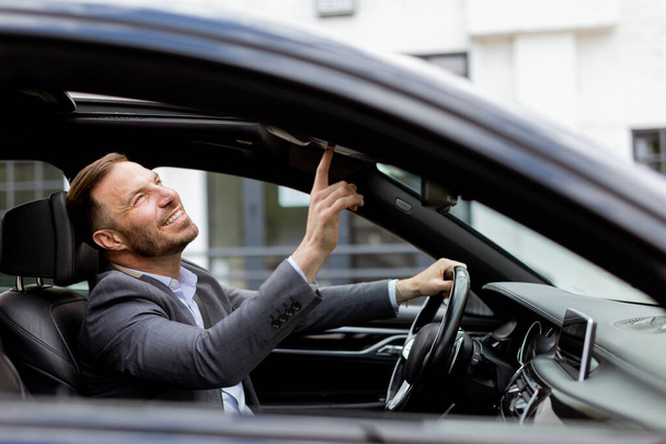 Usmívající se muž v obleku ovládá svůj kabriolet střešní mechanismus, zatímco parkuje na městské silnici - Fotografie, Obrázek