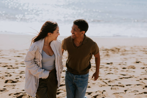 Una pareja sonriente disfruta de una carrera despreocupada en una orilla arenosa, olas suavemente rompiendo en el fondo - Foto, imagen