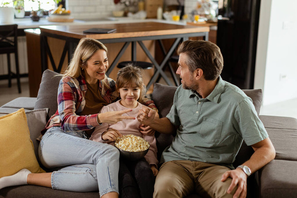 Egy vidám család üldögél a kanapén, játékos beszélgetést folytatva egy tál popcornnal. - Fotó, kép