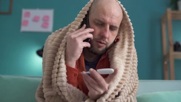 Family doctor concept. Unhappy bearded man suffering from flu holding a thermometer and making a call while calling to his doctor. Copy space - Footage, Video