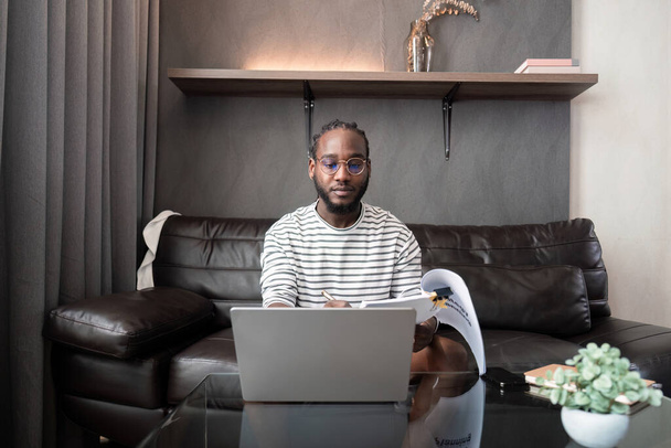 Afro-Amerikaanse man werkt met laptop computer afstandsbediening terwijl zitten op de bank in de woonkamer. Black guy doen freelance werk op kantoor. - Foto, afbeelding