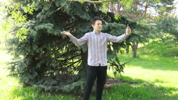 A young man stands in front of dense evergreen trees in a park, holding his phone at arms length to take a selfie. The sunlight filters through the foliage, highlighting the serene ambiance of this - Footage, Video