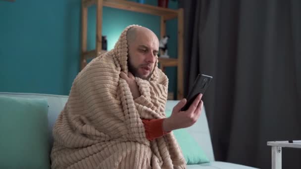 Człowiek z brodą chory w domu, używający smartfona do rozmowy z lekarzem przez wideokonferencję. Mężczyzna z objawami grypy z profesjonalnym lekarzem na czat online aplikacji. Przestrzeń kopiowania - Materiał filmowy, wideo