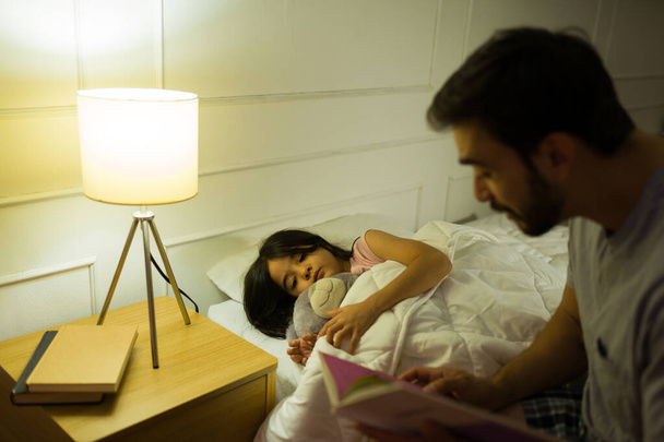 Isä lukee iltasadun nuorelle tyttärelleen, joka nukkuu sängyssä kotona lämpimällä, kodikas valaistus valaisee yön. - Valokuva, kuva