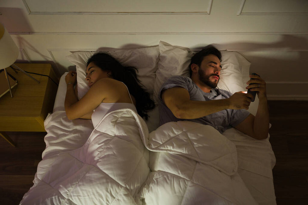 Jeune couple couché dans le lit la nuit, avec l'homme utilisant un smartphone pour éclairer son visage dans l'obscurité - Photo, image