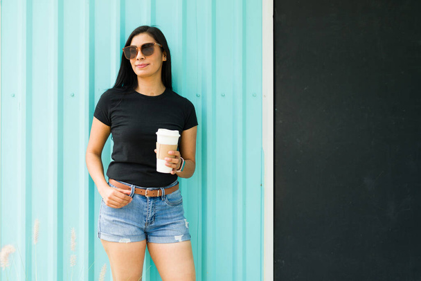 Pewna siebie latynoska kobieta w czarnej koszulce i okularach przeciwsłonecznych trzymająca filiżankę kawy na żywym turkusowym tle - Zdjęcie, obraz