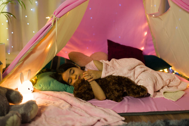 Küçük İspanyol kız, gece vakti peri ışıklarıyla süslenmiş ev yapımı çadırda pelüş hayvanıyla huzur içinde uyuyor. - Fotoğraf, Görsel