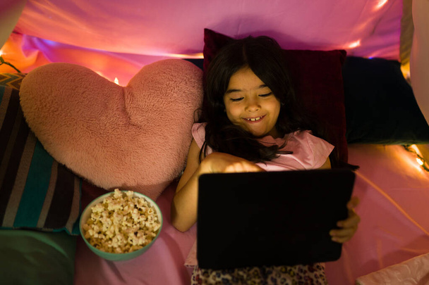 Uśmiechnięta latynoska dziewczynka leży w swojej kolorowej sypialni w nocy, używając tabletu do obejrzenia filmu i przekąski na popcornie - Zdjęcie, obraz