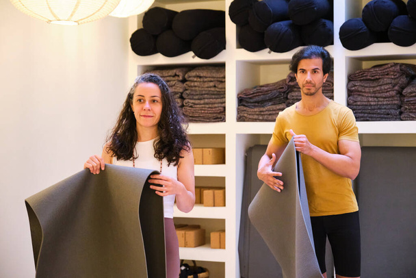 Два человека держат циновку yoga в комнате с полками полно материала yoga. Сцена спокойна и сосредоточена, так как люди, скорее всего, готовятся к занятию йогой. - Фото, изображение
