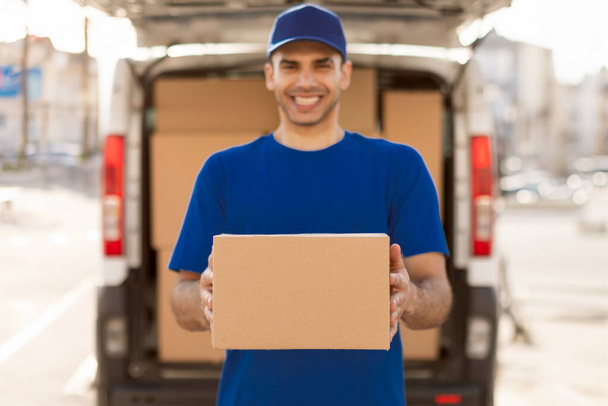 Homme joyeux transportant le paquet de la voiture de livraison, tenant la boîte en carton et souriant. Concept de service de messagerie - Photo, image