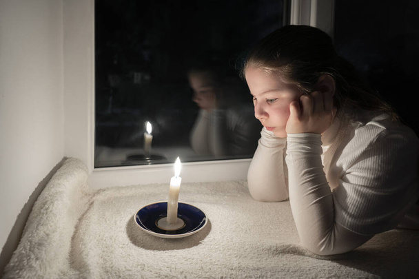 Blackout. A girl in a dark room lies on the windowsill near the window and looks at a burning candle. Power outage. Destruction of energy infrastructure - Photo, Image