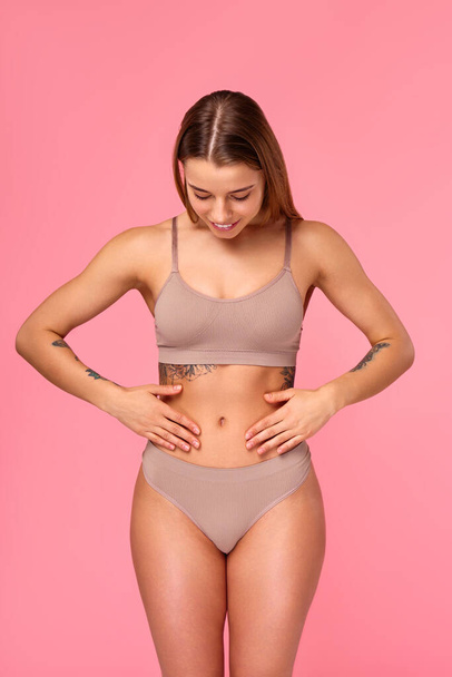 Mooie pasvorm vrouw in lingerie houden handen op de maag geïsoleerd op roze achtergrond. Lichaamsverzorging, kosmetologie, schoonheidsconcept. Sport en gewichtsverlies. - Foto, afbeelding
