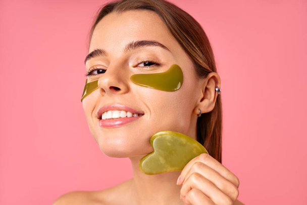 Beautiful woman with moisturizing patches under her eyes doing massage with gua sha scraper isolated on pink background. Facial skin care, cosmetology, beauty concept. - Photo, Image