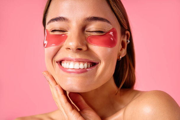 Belle femme avec des taches hydratantes sous ses yeux isolés sur fond rose. Soins du visage, cosmétologie, concept beauté. - Photo, image
