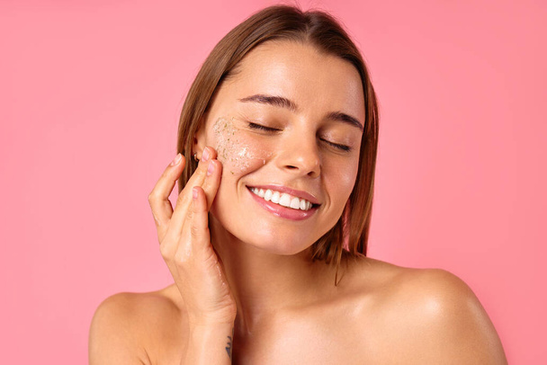 Beautiful happy woman using moisturizing face mask isolated on pink background. Skin care, cosmetology, beauty concept. - Photo, Image