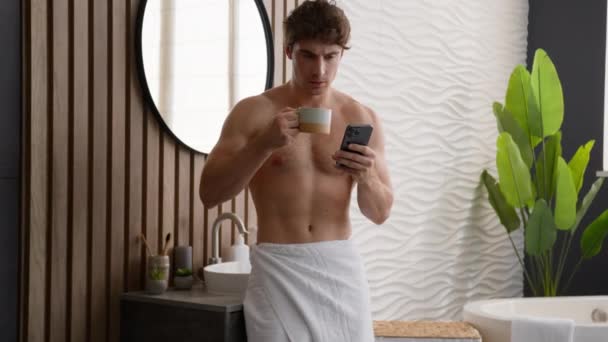 Valkoihoinen rento alaston mies juo teetä aamulla rutiini käsittelyssä uutisia sosiaalisen median älypuhelin selailu matkapuhelin juo kuumaa kahvia kylpyhuoneessa kaveri hämmästynyt shokki järkyttynyt sylkeminen juoma - Materiaali, video