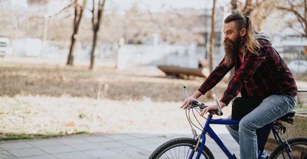 Homme d'affaires hippie en tenue décontractée utilisant un ordinateur portable assis sur un vélo bleu dans un parc urbain. - Photo, image