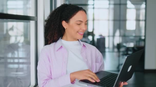 Mooie, succesvolle Braziliaanse of Spaanse krullende vrouw, software manager, bedrijfsmedewerker, staan in modern kantoor met een open laptop in handen, kijkt ernaar, glimlacht. Gebruik van gadgets en apparaten - Video