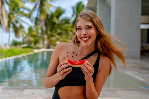 Radiante à beira da piscina, uma jovem beleza se aquece de felicidade de férias. Seu sorriso brilha enquanto ela devora saborosamente a melancia, simbolizando uma alusão despreocupada de verão.No encantador Caribe, uma mulher radiante faz uma pose em uma praia deslumbrante, sua silhueta g - Foto, Imagem