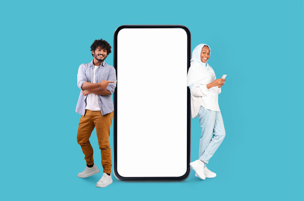 Hombre y mujer multirraciales en ropa relajada posando junto a una maqueta gigante de teléfonos inteligentes sobre un fondo azul - Foto, imagen