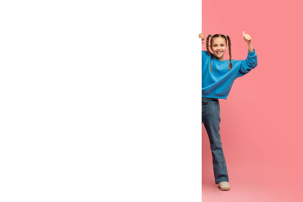 Ein fröhliches junges Mädchen in blauem Pullover und Jeans versteckt sich hinter einem leeren weißen Brett vor rosa Hintergrund, mit viel Kopierraum - Foto, Bild