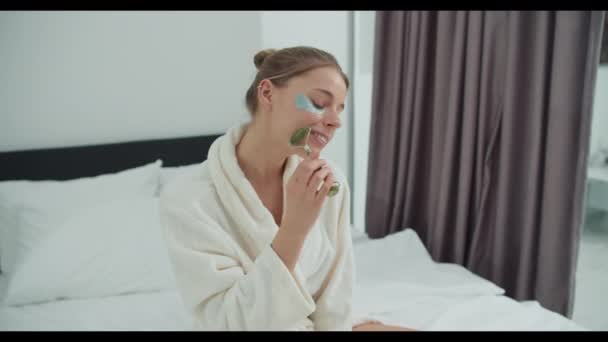 Een vrouw in een badjas zit op een bed met een roller op haar gezicht. Eyecatching gebaar intrigerend en leuk evenement - Video