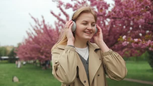 Retrato de la joven mujer caucásica feliz escuchando música en línea con auriculares inalámbricos desde un teléfono inteligente en la ciudad. Hermosa flor de cerezo sakura en primavera sobre el cielo azul. Sakura japonesa - Metraje, vídeo