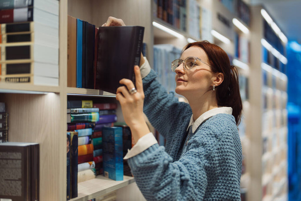 Gözlüklü odaklanmış genç bir kadın, iyi aydınlatılmış bir kütüphanedeki raftan bir kitap seçiyor. Farklı başlıkları keşfederken ilgisini ve merakını gösteriyor.. - Fotoğraf, Görsel