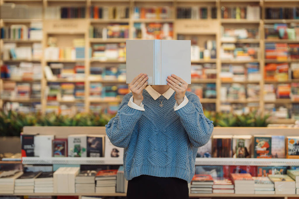 Una joven sostiene un libro frente a su portada, de pie en medio de una vibrante y variada librería. Estantes de libros crean un acogedor telón de fondo intelectual. - Foto, imagen